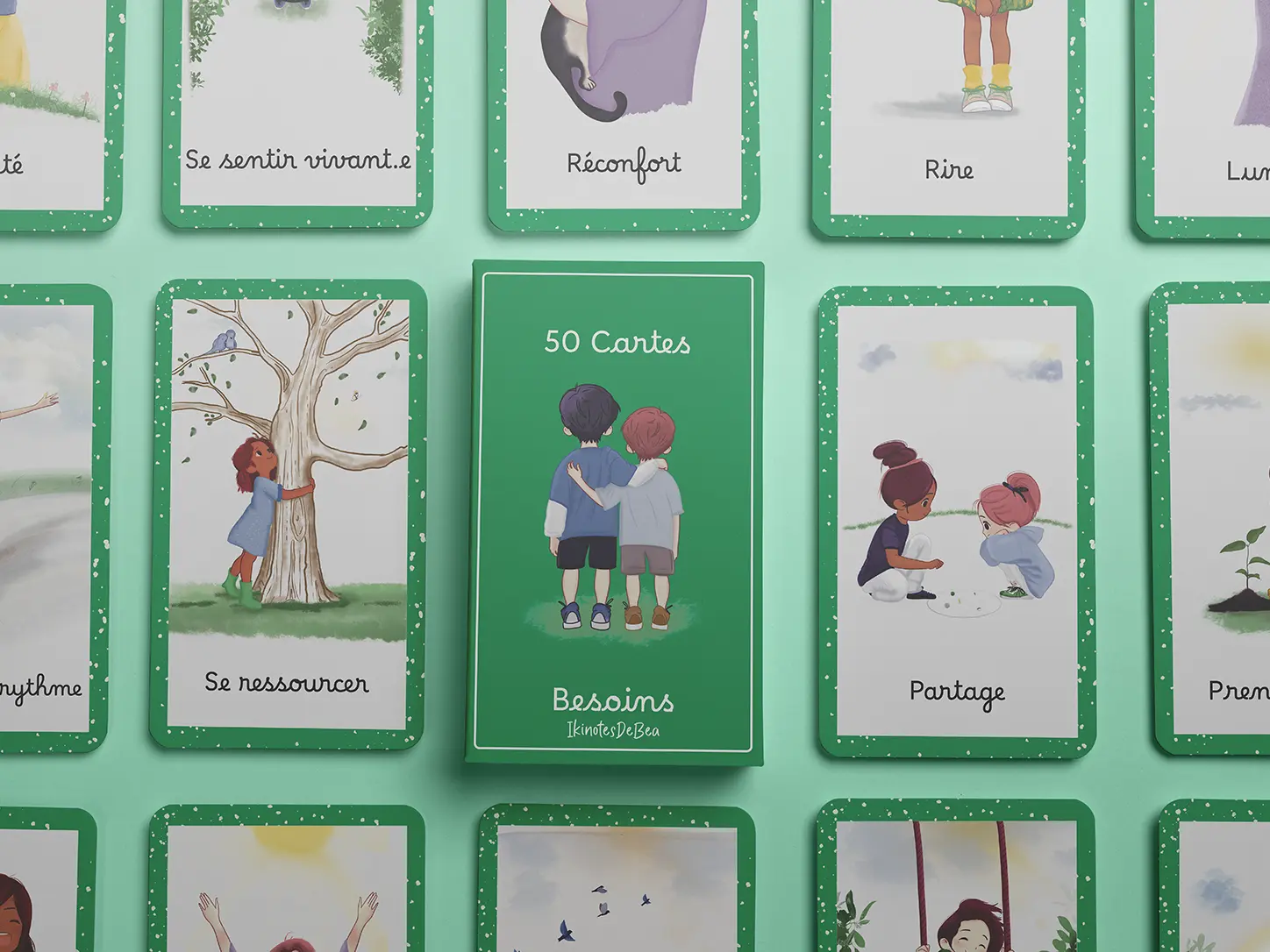 Maquette d'un jeu de 50 cartes «Besoins» avec boîtier vert et illustrations colorées d'enfants (se ressourcer, partage, réconfort, rire, se sentir vivant·e) disposées sur un fond vert pastel